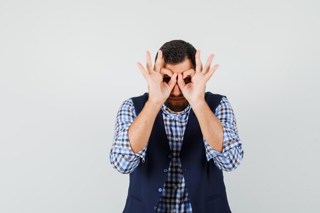 Young man showing glasses gesture in shirt, vest and looking focused