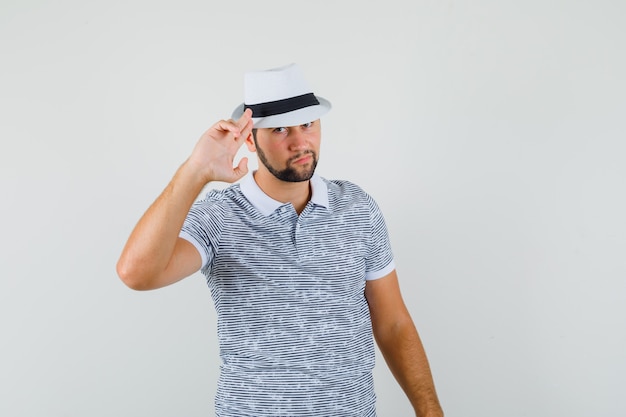 Молодой человек показывает жест прощания в футболке, шляпе и выглядит серьезным, вид спереди.
