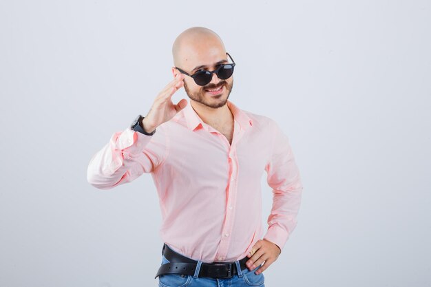 Young man showing greeting gesture in pink shirt,jeans,sunglasses and looking merry , front view.