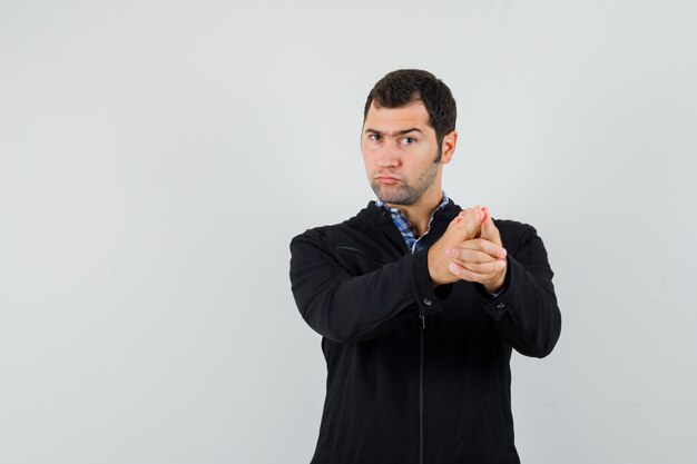 Молодой человек показывает жест пистолета в рубашке, куртке и выглядит сосредоточенным, вид спереди.