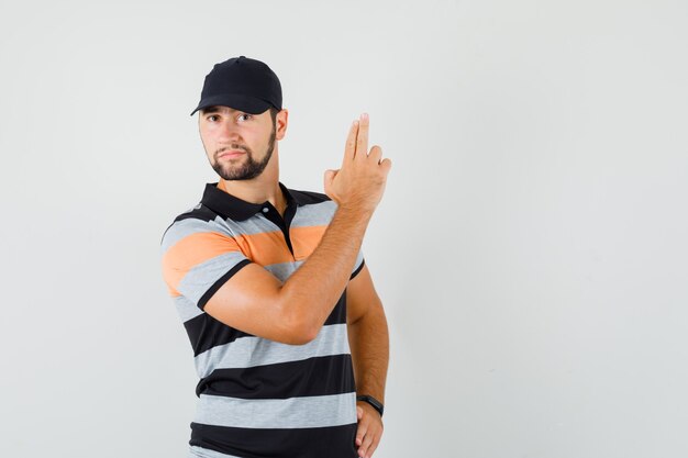 Young man showing gun gesture in t-shirt, cap and looking confident