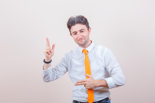 Young man showing gun gesture in white shirt, tie, jeans and looking confident