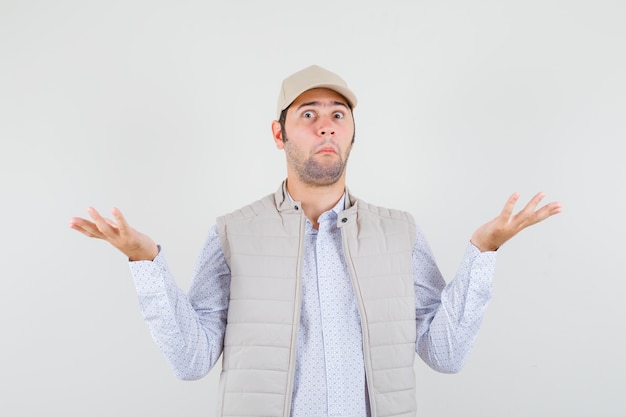 Young man showing helpless gesture in beige jacket and cap and looking surprised , front view.