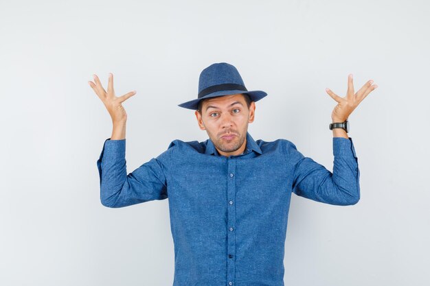 Young man showing helpless gesture in blue shirt, hat and looking puzzled , front view.