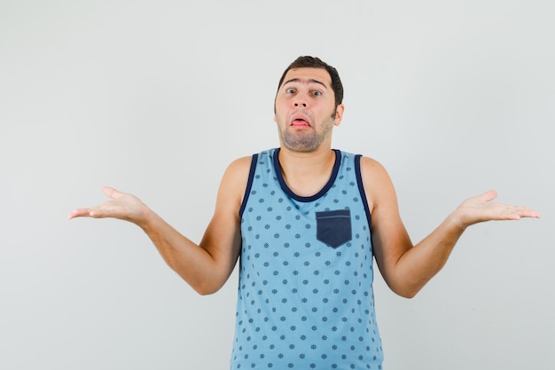 Young man showing helpless gesture in blue singlet and looking confused , front view.