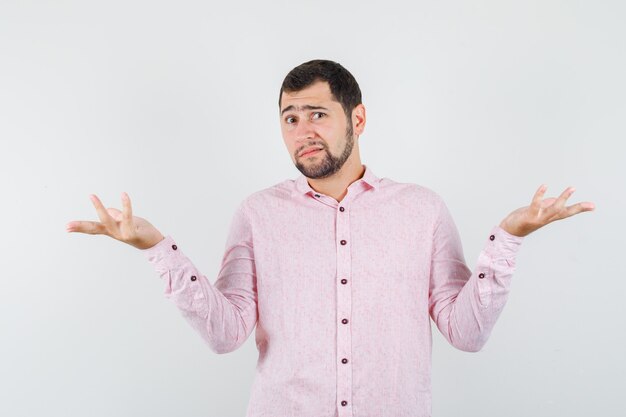 Young man showing helpless gesture in pink shirt and looking confused