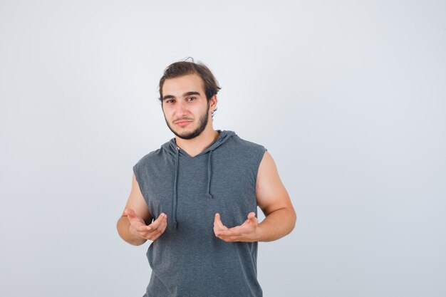 Young man showing helpless gesture in sleeveless hoodie and looking puzzled. front view.