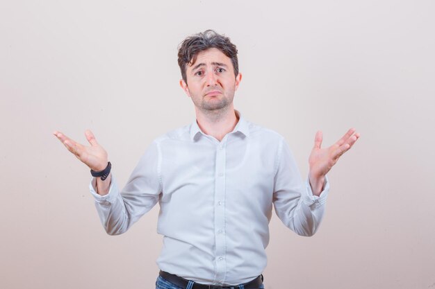 Young man showing helpless gesture in white shirt, jeans and looking confused