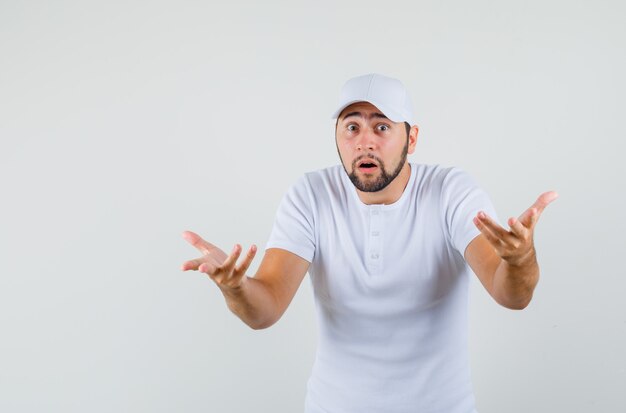 Young man showing helpless gesture in white t-shirt,cap and looking worried , front view.