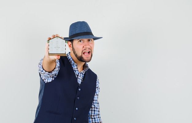 Young man showing house model in shirt, vest, hat