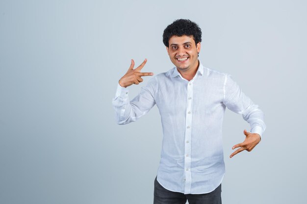 Young man showing I love you gesture in white shirt and looking merry , front view.