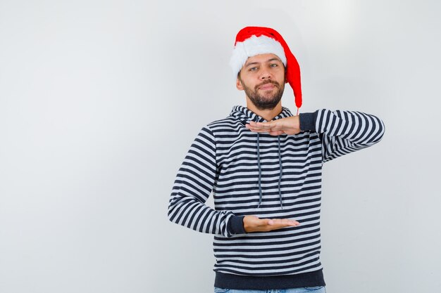 Young man showing large size sign in hoodie, Santa Claus hat and looking confident , front view.