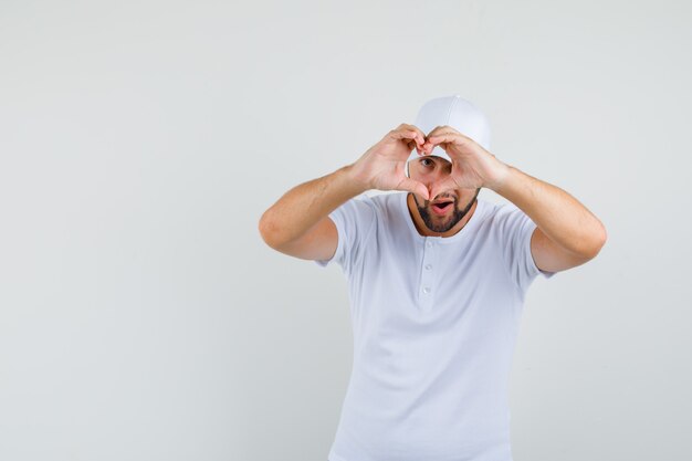 Young man showing love gesture in white t-shirt,cap and looking merry. front view. space for text