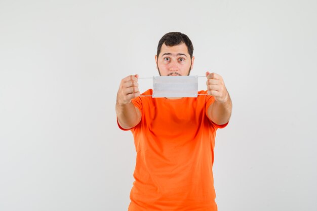 Young man showing medical mask in orange t-shirt and looking careful, front view.