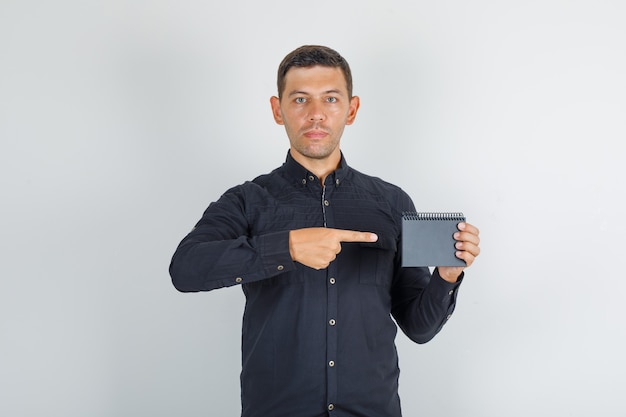 Young man showing mini notebook in black shirt, front view.