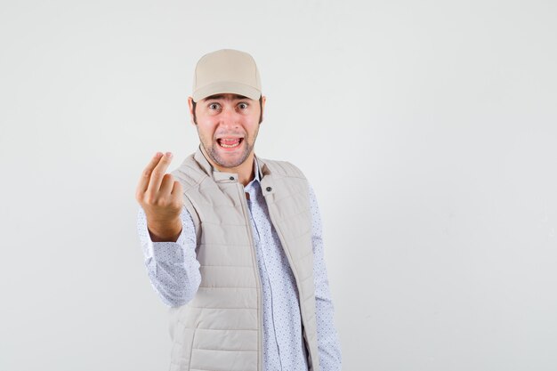 Молодой человек показывает денежный жест, высунув язык в бежевую куртку и кепку и выглядит счастливым. передний план.