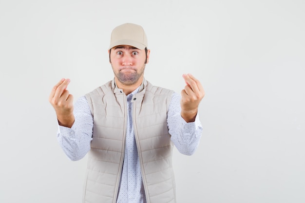 Young man showing money with both hands in beige jacket and cap and looking happy , front view.