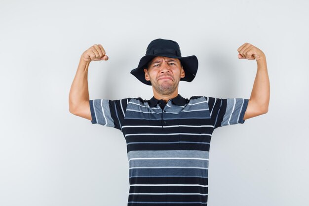 Young man showing muscles of arms in t-shirt, hat and looking strong. front view.