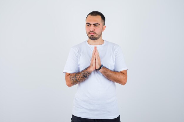 Young man showing namaste gesture in white t-shirt and black pants and looking pensive