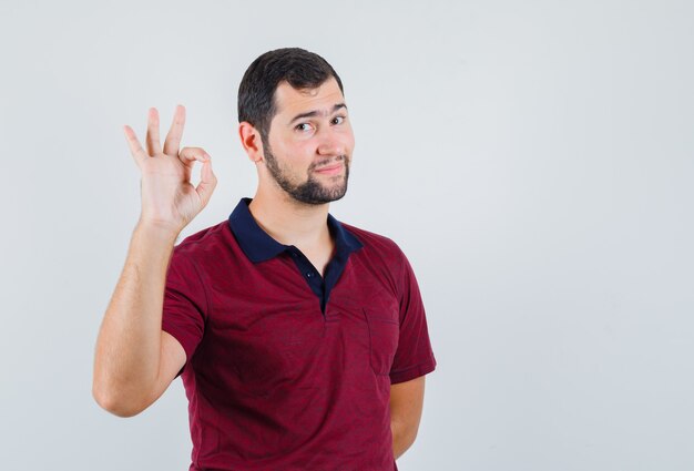 Young man showing ok gesture in red t-shirt and looking pleased. front view.