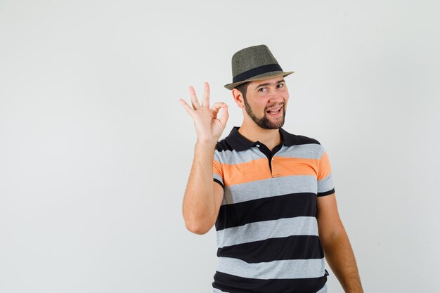 Молодой человек показывает жест в футболке, шляпе и выглядит весело