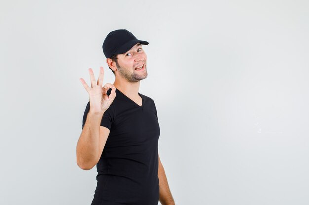 Young man showing ok sign in black t-shirt, cap and looking cheerful.