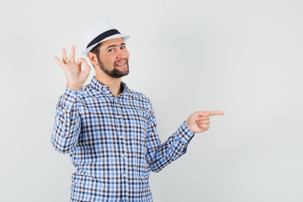 Молодой человек показывает знак ОК, указывая в сторону в клетчатой рубашке, шляпе и выглядит весело