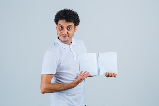 Young man showing opened notebook in white t-shirt and jeans and looking serious , front view.