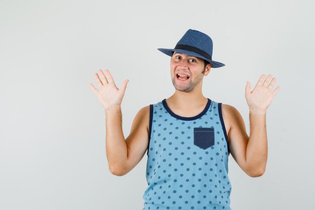 Young man showing palms in surrender gesture in blue singlet, hat and looking glad. front view.