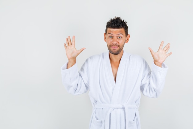 Young man showing palms in surrender gesture in white bathrobe and looking positive. front view.