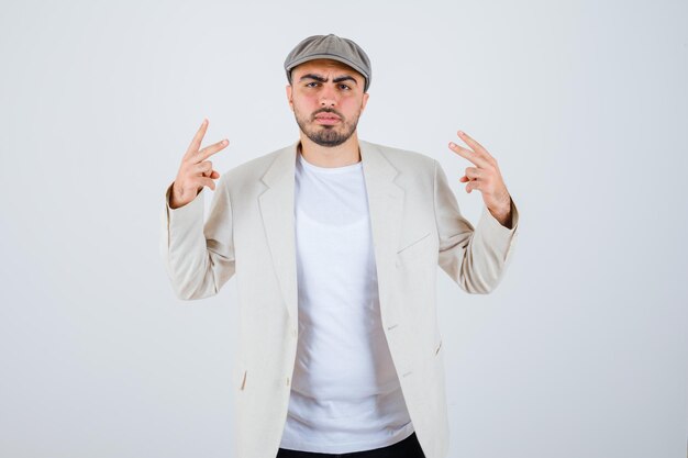 Young man showing peace signs in white t-shirt, jacket and grey cap and looking angry