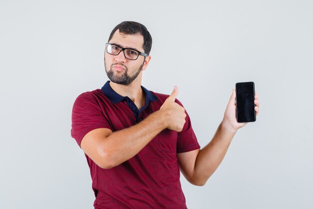 Young man showing phone while thumb up in red t-shirt,optic glasses and looking pleased. front view.