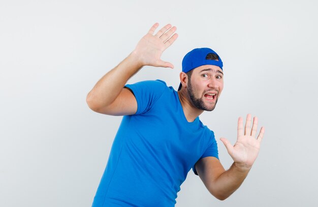 Young man showing refusal gesture in blue t-shirt and cap and looking frightened