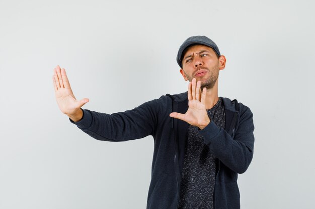 Young man showing refusal gesture to protect himself in t-shirt, jacket, cap