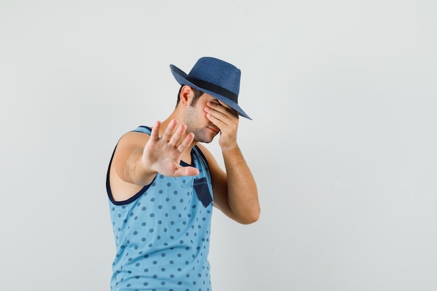 Young man showing refusal gesture with hand on eyes in blue singlet, hat and looking scared , front view.