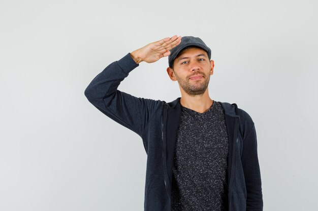 Young man showing salute gesture in t-shirt, jacket, cap and looking confident.