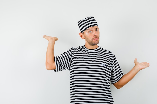 Free photo young man showing scales gesture in striped t-shirt, hat and looking irresolute.