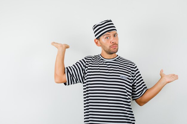 Young man showing scales gesture in striped t-shirt, hat and looking irresolute.