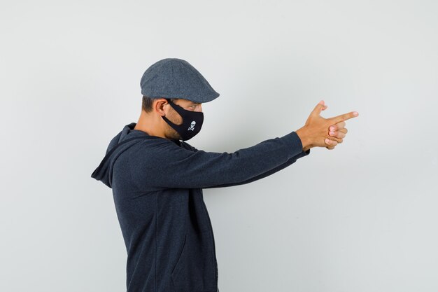 Young man showing shooting gesture in t-shirt, jacket, cap, mask and looking focused. .