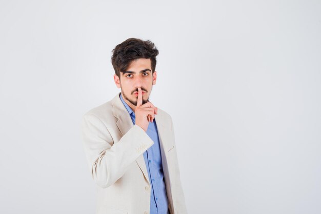 Young man showing silence gesture in blue t-shirt and white suit jacket and looking serious