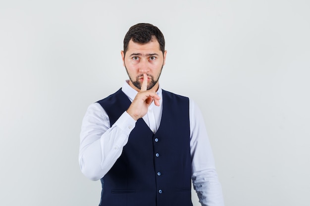 Young man showing silence gesture in shirt and vest and looking careful