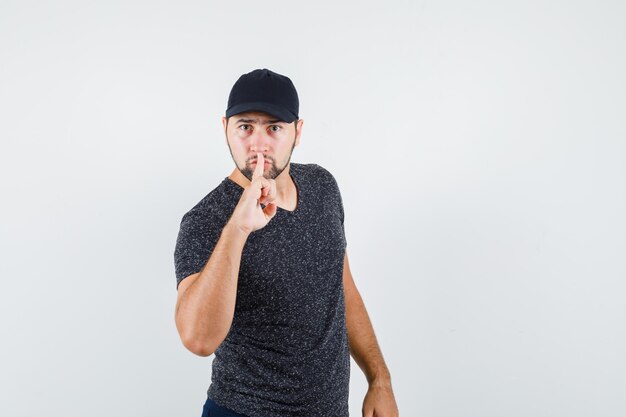 Young man showing silence gesture in t-shirt and cap and looking serious