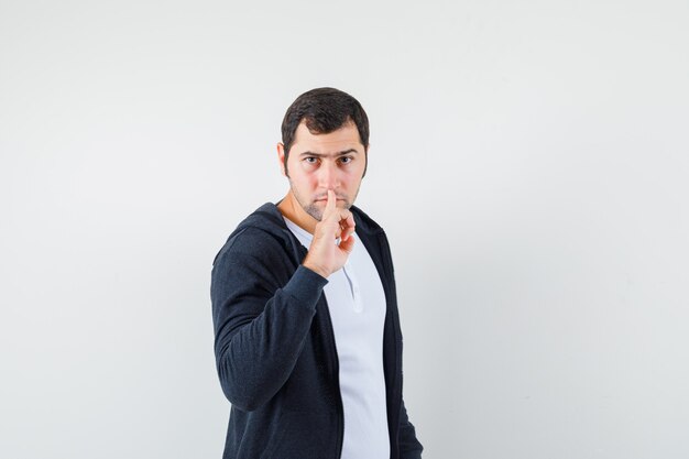 Young man showing silence gesture in white t-shirt and zip-front black hoodie and looking serious. front view.
