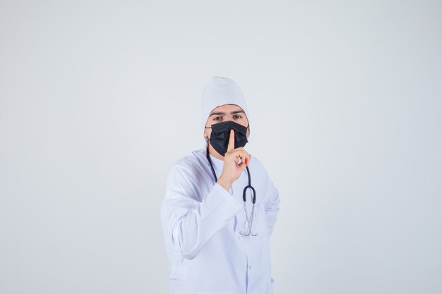 Young man showing silence gesture in white uniform, mask and looking confident. front view.