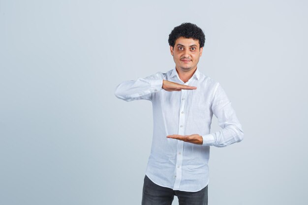 Young man showing size sign in white shirt, pants and looking confident , front view.