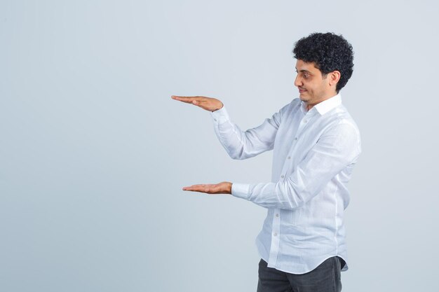 Young man showing size sign in white shirt, pants and looking focused. front view.