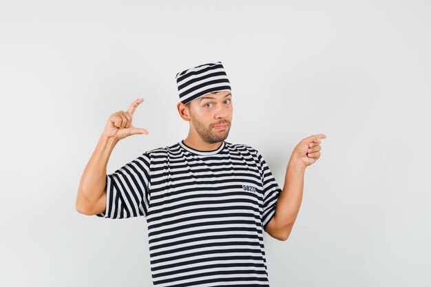 Young man showing small size sign, pointing aside in striped t-shirt, hat.