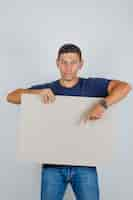 Free photo young man showing something on poster in blue t-shirt, jeans and looking positive, front view.