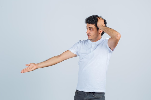 Young man showing something below in white t-shirt, pants and looking confused. front view.