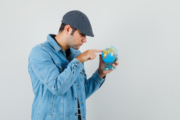 Young man showing somewhere on mini globe in jacket,cap and looking concentrated. front view.